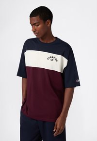 Młody mężczyzna w kolorowej koszulce Champion w granatowym, białym i burgundowym kolorze, z rękami w kieszeniach, patrzący w dół na jednolitym tle.