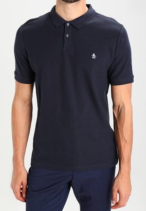 Homme portant un polo bleu marine à manches courtes avec un petit logo pingouin blanc sur la poitrine gauche, associé à un pantalon bleu marine.