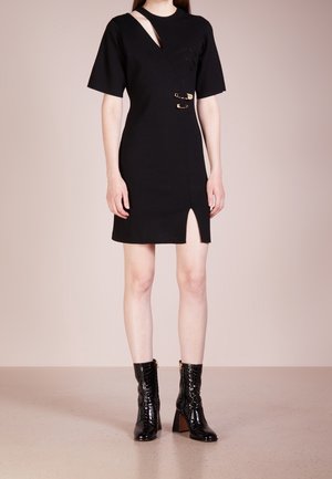 Kobieta ubrana w czarną sukienkę z krótkim rękawem, z wykrojonym dekoltem, złotymi detalami w formie agrafek, rozcięciem z przodu oraz czarne botki na obcasie do połowy łydki.