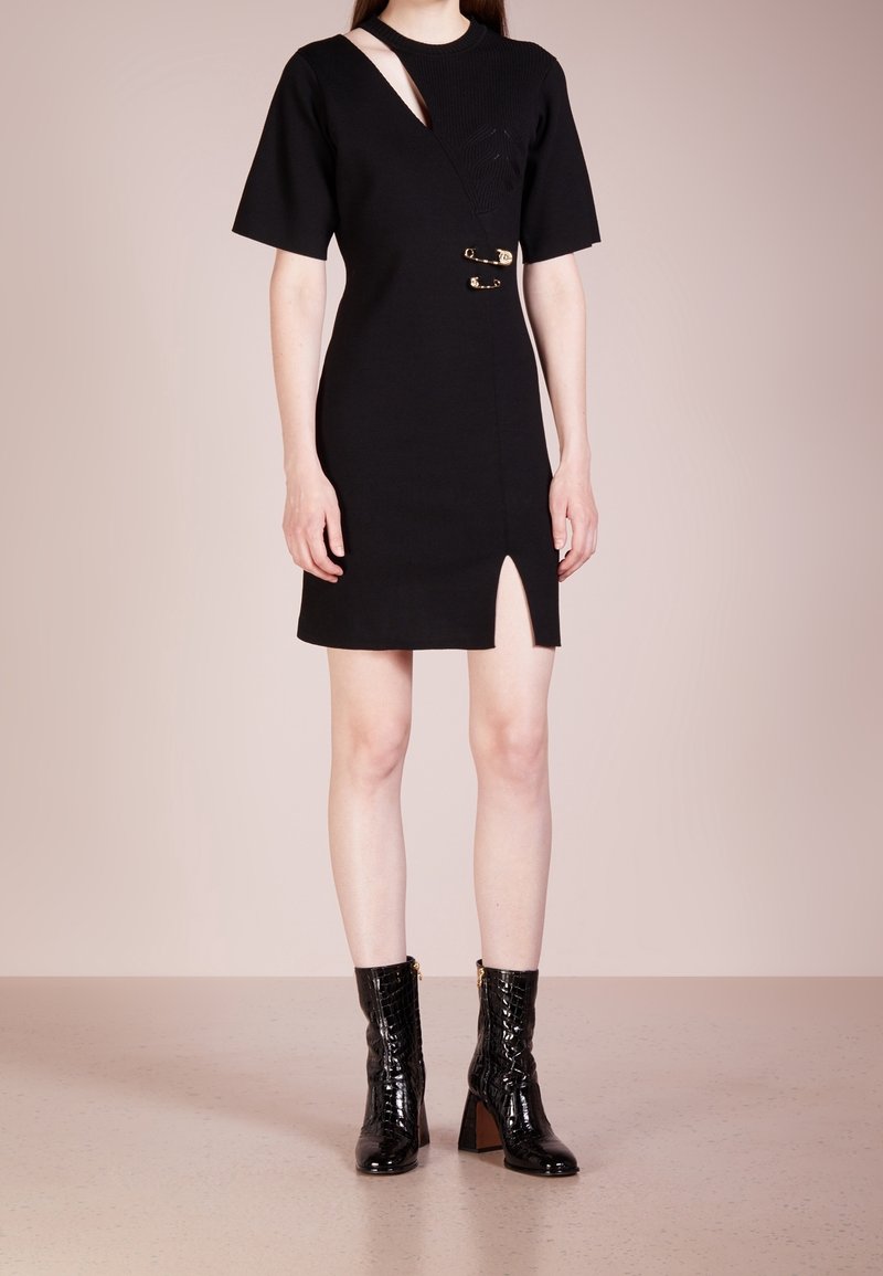 Femme portant une robe noire à manches courtes avec un décolleté découpé, des détails en épingles de sûreté dorées, une fente frontale, et des bottes noires à talons mi-mollet.