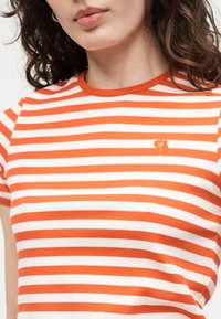 Γυναίκα που φοράει μπλούζα με πορτοκαλί και λευκές ρίγες, με μικρή πορτοκαλί κεντημένη αρκούδα στο στήθος, δείχνοντας τον λαιμό και τα σγουρά μαλλιά της.