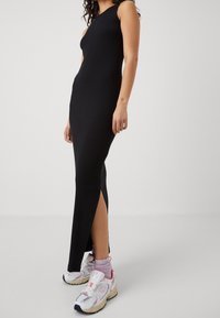 Černé maxi šaty bez rukávů s bočním rozparkem; hladká, pružná látka. Kombinované s bílými a růžovými teniskami a ponožkami s růžovými akcenty.