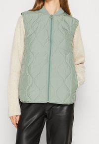 Jasnozielona, pikowana kamizelka z wysokim kołnierzem, zapinana z przodu na zamek błyskawiczny, z tonalnym przeszyciem, w połączeniu z kremowym swetrem i czarnymi spodniami.