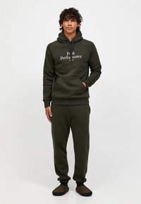 Grüne Kapuzenjacke mit "Peak Performance"-Logo, Kängurutasche und Kordeln; passende Jogginghosen; Modell trägt braune Slipper.