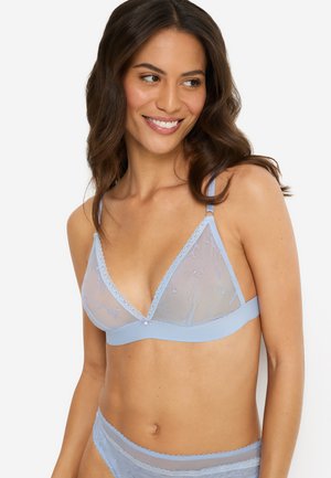 Χαμογελαστή γυναίκα που φοράει ανοιχτό μπλε δαντελένιο εσώρουχο με κεντήματα λουλουδιών, κοιτάζοντας ελαφρώς προς τα πλάγια σε ένα απλό λευκό φόντο.