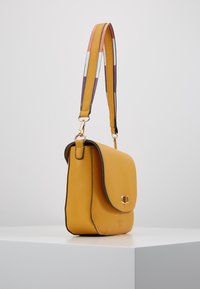 Žlutá kožená kabelka na rameno s klopou, kontrastním černým lemem a ozdobným vícebarevným pruhovaným popruhem. Akcenty ze zlaté kovových doplňků.