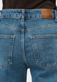 Πίσω όψη ατόμου που φοράει τζιν παντελόνι με ψηλή μέση, δύο πίσω τσέπες και ένα ταμπελάκι από ταμπά δέρμα στη μέση.