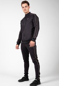 Zwarte sportieve trainingspak met gouden zijstreep. De jas heeft een rits aan de voorkant, een hoge kraag en aansluitende manchetten. De broek heeft een aansluitende pasvorm.