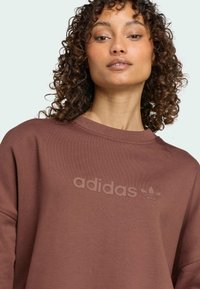 Hnědá mikina vyrobená z měkké látky s kulatým výstřihem, zdobená decentním logem adidas na hrudi a volným střihem.