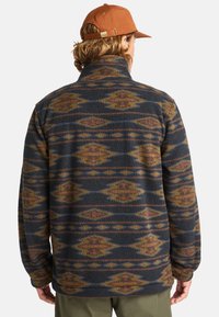 Pull en polaire avec un col haut, arborant un motif géométrique dans des tons terreux de marron, jaune et bleu sur un fond sombre.