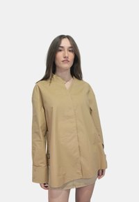 Camicia a maniche lunghe beige con colletto alla cinese, realizzata in tessuto leggero. Presenta polsini con bottone e una vestibilità rilassata con orlo curvo.