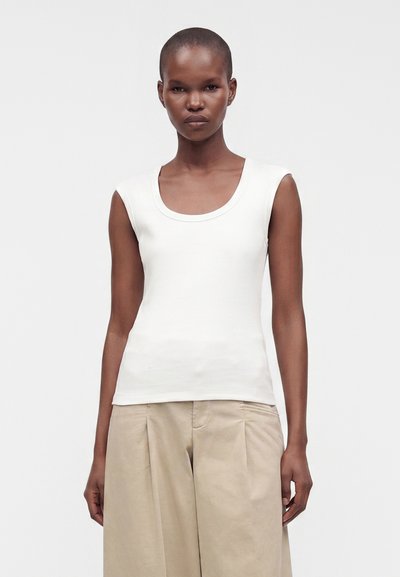 Femme aux cheveux courts portant un haut blanc sans manches et un pantalon large beige, debout devant un fond clair uni.