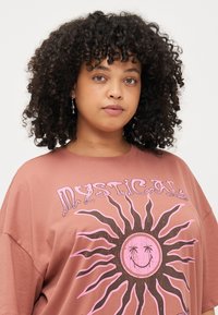 Camiseta oversize marrón con un diseño gráfico de sol, con acentos en rosa y una cara sonriente. Mangas cortas y textura suave y lisa.