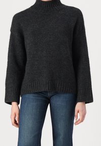 Personne portant un pull à col roulé côtelé gris foncé avec des manches larges et un jean en denim bleu sur un fond uni.