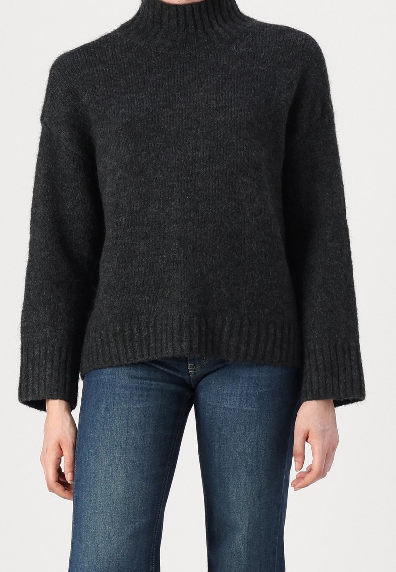 Personne portant un pull à col roulé côtelé gris foncé avec des manches larges et un jean en denim bleu sur un fond uni.