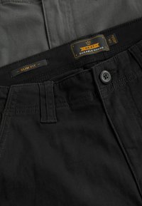 Schwarze slim-fit Hosen mit glatter Textur, einem Knopfverschluss und einem Etikett, das "NXT Durable Goods" sowie zusätzliche Größenangaben enthält.