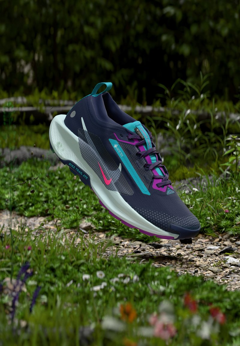 Πέδιλο τρεξίματος σε μονοπάτια της Nike σε σκούρο μπλε και μοβ με πράσινες λεπτομέρειες, ετικέτα GORE-TEX και μαξιλαρωμένη λευκή σόλα που αιωρείται πάνω από μονοπάτι με χαλίκι.