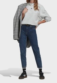 Grå sweatshirt med broderad logotyp, högmidjade blå jeans, svarta sneakers med vita ränder, och en grå rutiga kavaj över axeln.