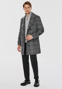 Szary, kraciasty płaszcz o podwójnym zapięciu, połączony z jasnoszarym, zapinanym na zamek kołnierzykowym swetrem, czarnymi spodniami i błyszczącymi czarnymi butami.