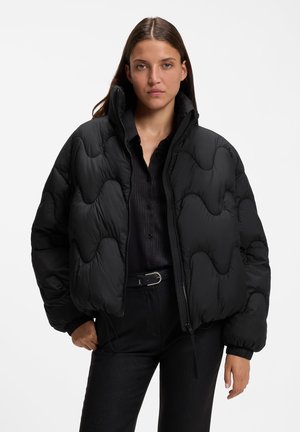 Frau trägt eine schwarze gesteppte Daunenjacke über einem schwarzen Hemd und einer schwarzen Hose, steht mit neutralem Gesichtsausdruck vor einem schlichten Hintergrund.