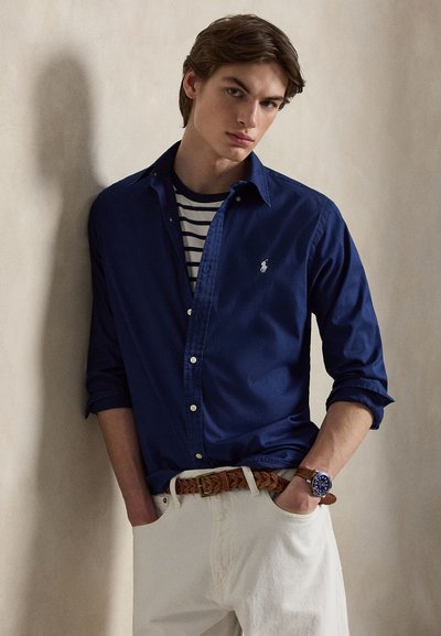 Νεαρός άνδρας με μπλε πουκάμισο με κουμπιά πάνω από ριγέ μπλουζάκι, λευκό παντελόνι, καφέ πλεκτή ζώνη και μπλε ρολόι χειρός, στέκεται με τα χέρια στις τσέπες.