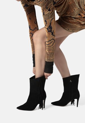 Stivaletti in suede neri con punta a cono e tacco a stiletto. I dettagli includono una rifinitura frangiata nella parte superiore e una cerniera laterale.