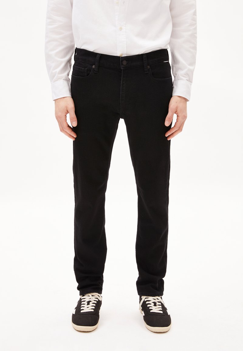 Jean en denim noir avec une coupe slim, présentant des poches avant, des passants de ceinture et une fermeture à bouton. Associé avec des baskets noires.