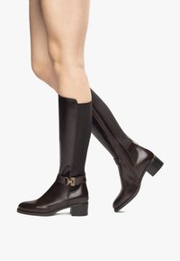 Braune kniehohe Lederstiefel mit seitlichem Reißverschluss, elastischem Einsatz und einer kleinen dekorativen Schnalle. Glossy Finish und Blockabsatz.