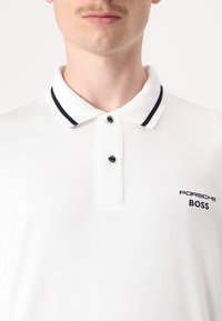 Balta polo marškiniai su applikuota apykakle, turinčia juodas juosteles, dviejų mygtukų užsegimą ir siuvinėtą "PORSCHE BOSS" logotipą ant krūtinės.