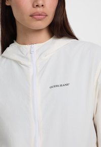 Balta gobtuvė su užtrauktuku priekyje ir subtiliu tekstūru. Kairėje pusėje yra juodas "GUESS JEANS" logotipas.