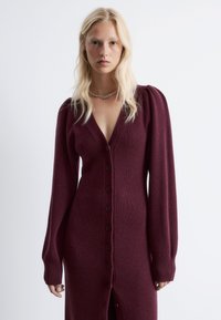 Burgunderfarbener Strickcardigan-Kleid mit V-Ausschnitt, langen Puffärmeln, durchgehender Knopfleiste und gerippten Bündchen. Weiche Textur und schlitz bis zur Oberschenkelhöhe.