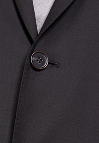 Svart skräddarsydd jacka med en slät textur, skarp slag och en enkel rund knapp med fyra hål, mot en ljusgrå bakgrund.