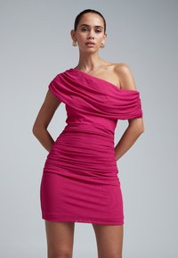 Moteris vilki ryškiai rožinę vieno peties raukšlėtą mini suknelę su tekstūruotais auksiniais auskarais, stovi priešais paprastą pilką foną.