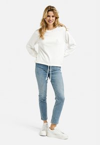 White sweatshirt with drawstring hem, featuring a small pink graphic on the chest, paired with light blue denim jeans and white sneakers.