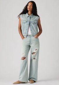 Femme portant une veste en jean sans manches bleu clair, un haut blanc, un jean large déchiré et des ballerines beige, debout les mains dans les poches.