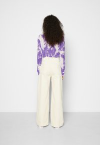 Camisa de manga longa em tie-dye roxo, calças largas em creme, bolsos traseiros visíveis e leve textura no tecido. Design simples e moderno.