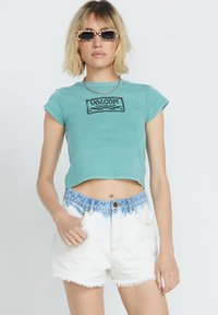 Turquoise crop top met korte mouwen en een zwart grafisch ontwerp. Gecombineerd met witte denim shorts met een lichtblauwe ombre tailleband.