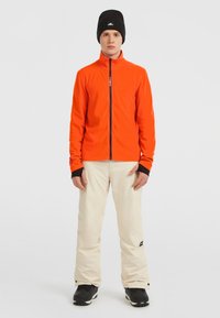 Heldere oranje zip-up jack met zwarte accenten, gecombineerd met crèmekleurige skibroeken. Het model draagt een zwarte beanie en zwarte skischoenen.