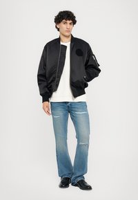 Μαύρο bomber jacket με λεία υφή και κυκλικό λογότυπο, συνδυασμένο με λευκό πουλόβερ και φθαρμένα ανοιχτόχρωμα μπλε τζιν.