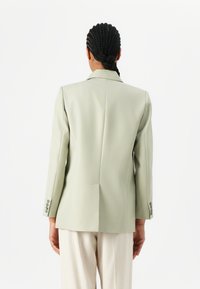 Lys grønn skreddersydd blazer med en enkelt lufteventil, strukturerte skuldre og sølvfargede knappdetaljer. Stylet med lyse bukser.