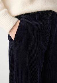 Personne portant un pantalon en velours côtelé noir avec une main dans la poche et une manche de pull en peluche de couleur crème visible.