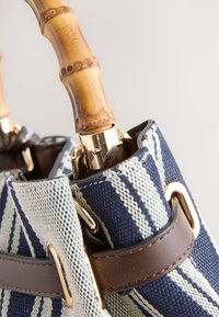 Sac en tissu rayé bleu et blanc avec une sangle en cuir marron et une poignée incurvée en bambou fixée par une quincaillerie dorée.