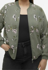 Grøn bomberjakke med blomsterprint, lynlås lukning, ribbede manchetter og kant; bæres over en sort top, suppleret med højtaljede sorte jeans.