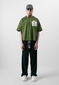 Grønn avkortet T-skjorte med en frontlapp og korte ermer. Kombinert med løse svarte bukser og hvite sneakers. Grå caps fullfører looken.