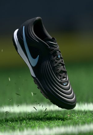 Czarny korki piłkarskie Nike z jasnoniebieskim logo w powietrzu nad zielonym boisku z białą linią.