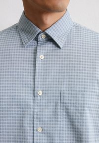 Light blue checkered button-up shirt with a spread collar, white buttons, and a single chest pocket. Fabric has a textured finish.