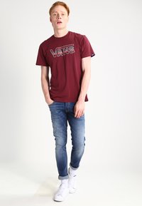 Crvena pametna majica s logotipom "VANS", uparen s svijetloplavim trapericama i bijelim tenisicama. Standardni kroj s kratkim rukavima i okruglim izrezom.