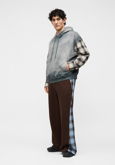 Άνδρας στέκεται με τα χέρια στις τσέπες, φορά γκρι φούτερ με κουκούλα και καρό μανίκια και καφέ παντελόνι με μπλε καρό πλαϊνές ρίγες.