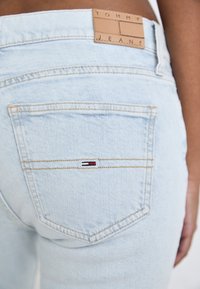 Vaqueros de denim de lavado ligero con una etiqueta de cuero marrón que dice "TOMMY JEANS". El bolsillo trasero tiene un pequeño detalle de logotipo en rojo, blanco y azul.