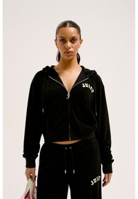 Tuta in velluto nero con cappuccio zip e tasca frontale, con la scritta "JUICY" in verde. Pantaloni abbinati con vita regolabile.